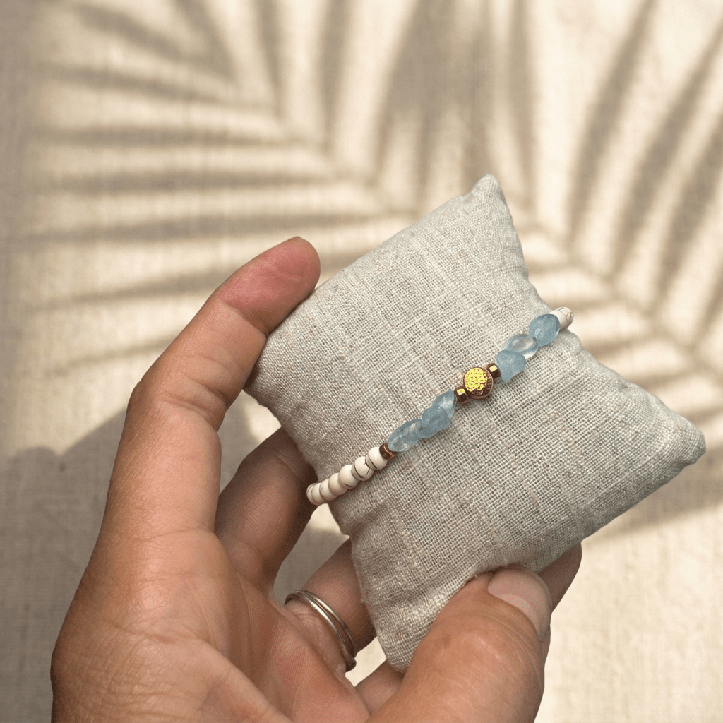 Handgemachtes Aquamarin Armband mit weiß-beigen Perlen und goldener Blume-des-Lebens-Perle auf einem Leinenkissen.