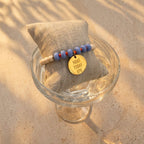 Blaues Perlenarmband mit goldenem Plättchen auf Glasschale im Sand – Mantra Schmuck für Damen