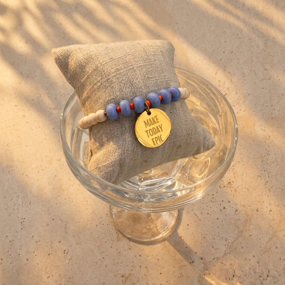 Blaues Perlenarmband mit goldenem Plättchen auf Glasschale im Sand – Mantra Schmuck für Damen