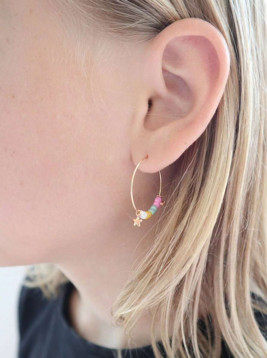Goldene Hoop-Creolen mit bunten Heishi-Perlen in Regenbogenfarben und goldenem Stern-Anhänger, getragen von einem Mädchen mit blonden Haaren. Trueffeldesign
