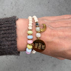Buntes Perlenarmband Gold mit goldenem Mantra-Anhänger am Handgelenk, kombiniert mit grauem Strickpullover
