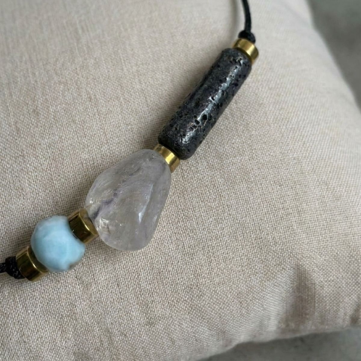 Nahaufnahme eines eleganten Diffuser Armbands mit hellblauem Larimar, klarem Rutilquarz und schwarzem Lavastein auf einem Leinenkissen.