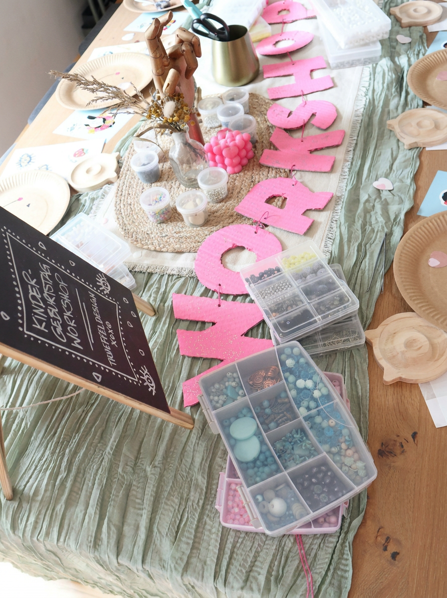 Kinder-Geburtstag Schmuck-Workshop Tisch von oben mit pinkem WORKSHOP-Schriftzug, Perlensortierkästen, Holztabletts, Tafelaufsteller und grünem Musselin-Tischläufer. Trueffeldesign