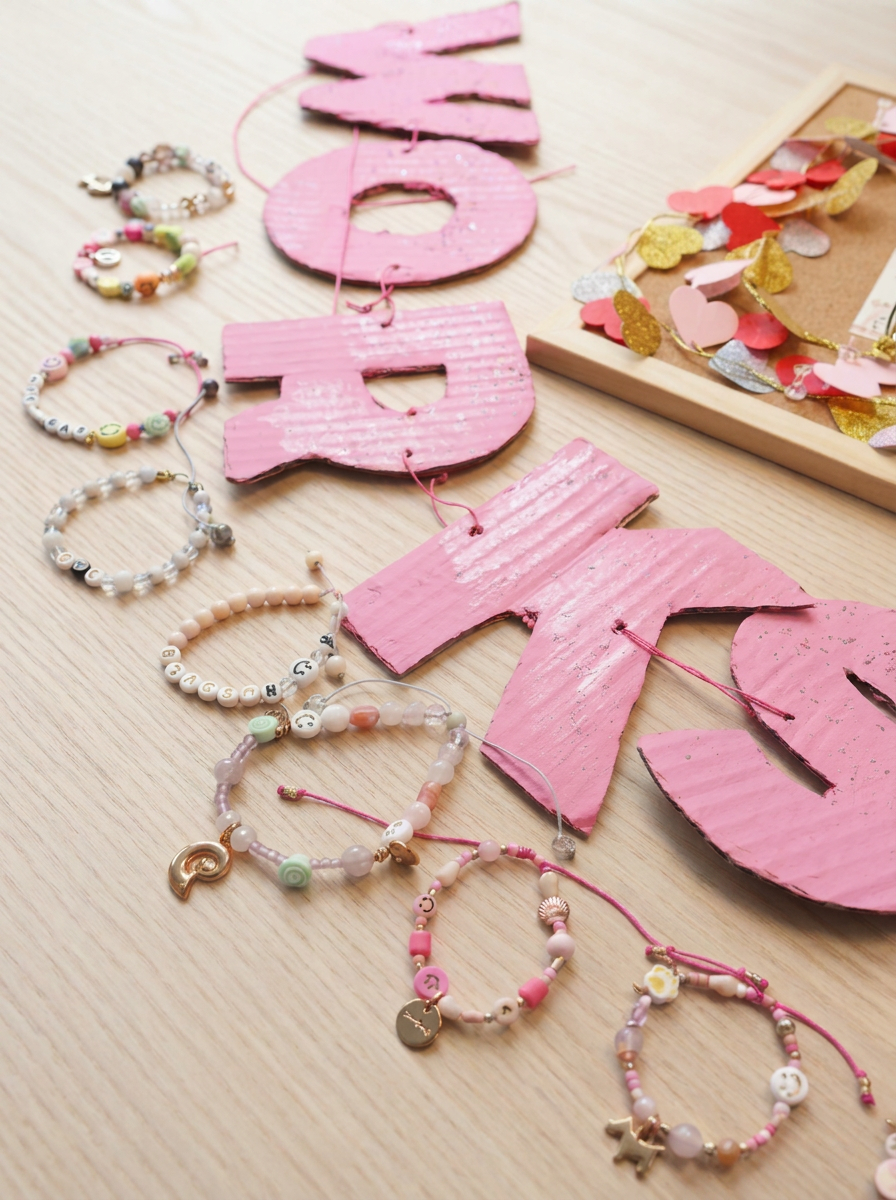 Fertige selbstgemachte Kinder-Perlenarmbänder in verschiedenen Styles – mit Buchstabenperlen, Edelsteinperlen, Heishi-Perlen, Charms und Smileys neben pinkfarbenem WORKSHOP-Schriftzug. Trueffeldesign
