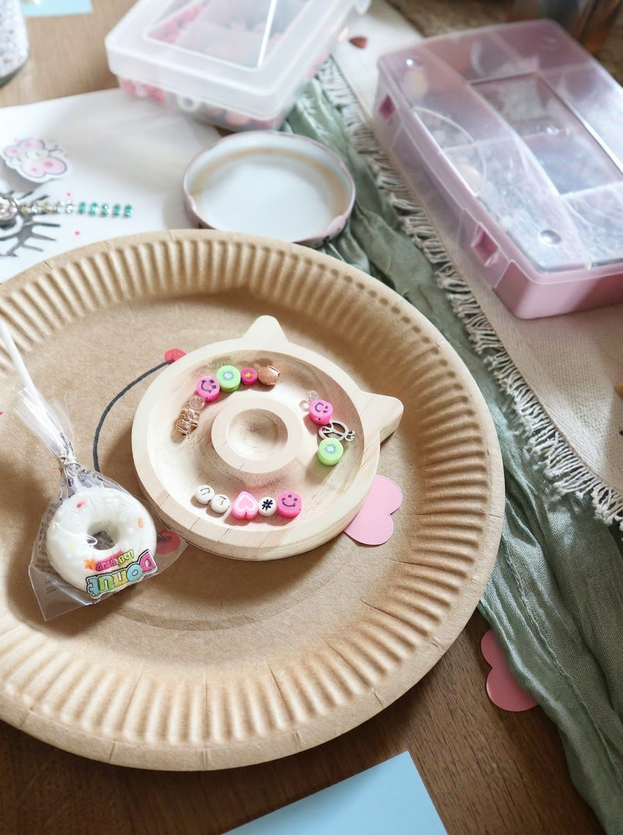 Kinder Schmuck Workshop Setup mit rundem Holz-Perlentablett in Katzenform, bunten Perlen, Buchstabenperlen und Donut-Süßigkeit auf Pappteller. Trueffeldesign