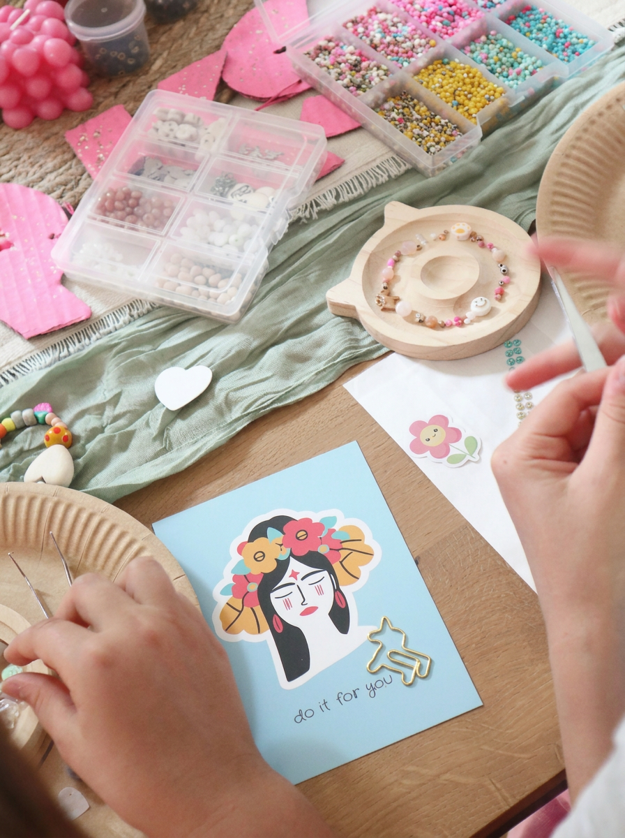 Kinder-Schmuck-Workshop-Tisch mit hellblauer Frida-Kahlo-Postkarte mit Aufschrift do it for you, bunten Miyuki-Perlen in Sortierbox, Holz-Armband-Tablett und pinkem Workshop-Schriftzug. Trueffeldesign