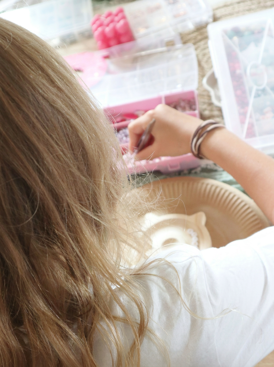 Mädchen mit langen blonden Haaren und Armbändern sucht konzentriert Perlen aus pinkfarbener Sortierbox beim Kinder-Schmuck-Workshop. Trueffeldesign