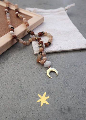 Mala Kette aus warmem Sonnenstein, Sandelholz und einem goldenen Mondanhänger aus Edelstahl neben einem Säckchen.