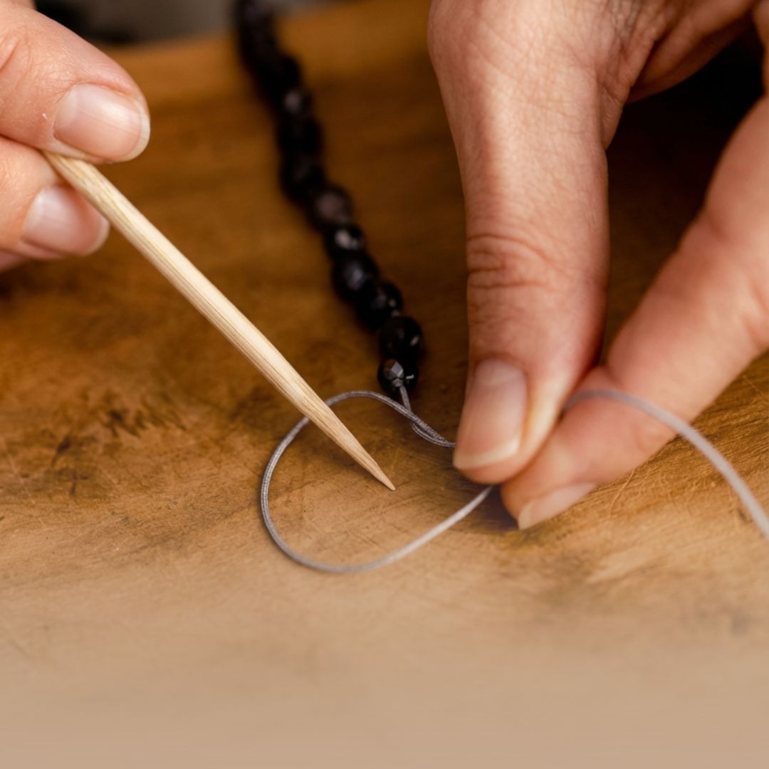 Nahaufnahme von Händen, die unter fachkundiger Anleitung eine Mala-Kette mit der traditionellen Knüpftechnik fertigen.