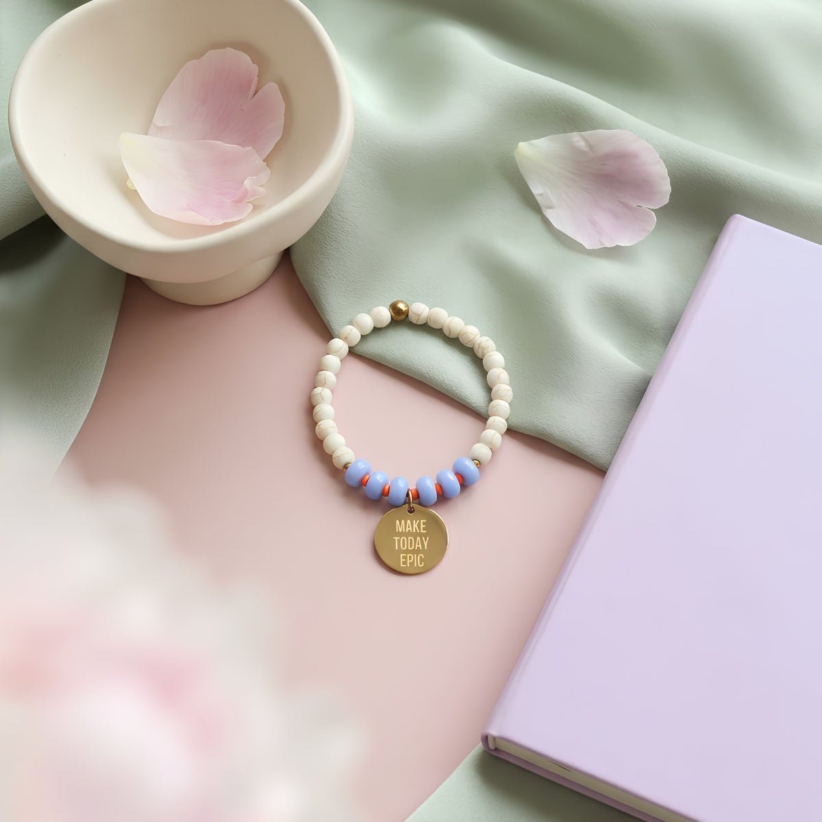 Mantra Schmuck Damen – blaues Perlenarmband mit goldenem Plättchen auf pastellem Flatlay mit Rosenblüten
