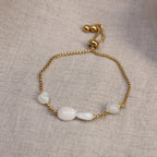 Flach liegendes Damen-Armband aus vergoldetem Edelstahl mit echten, weiß schimmernden Mondstein-Perlen und Zugverschluss auf Leinenstoff.