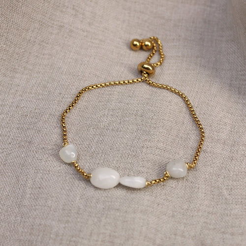 Flach liegendes Damen-Armband aus vergoldetem Edelstahl mit echten, weiß schimmernden Mondstein-Perlen und Zugverschluss auf Leinenstoff.