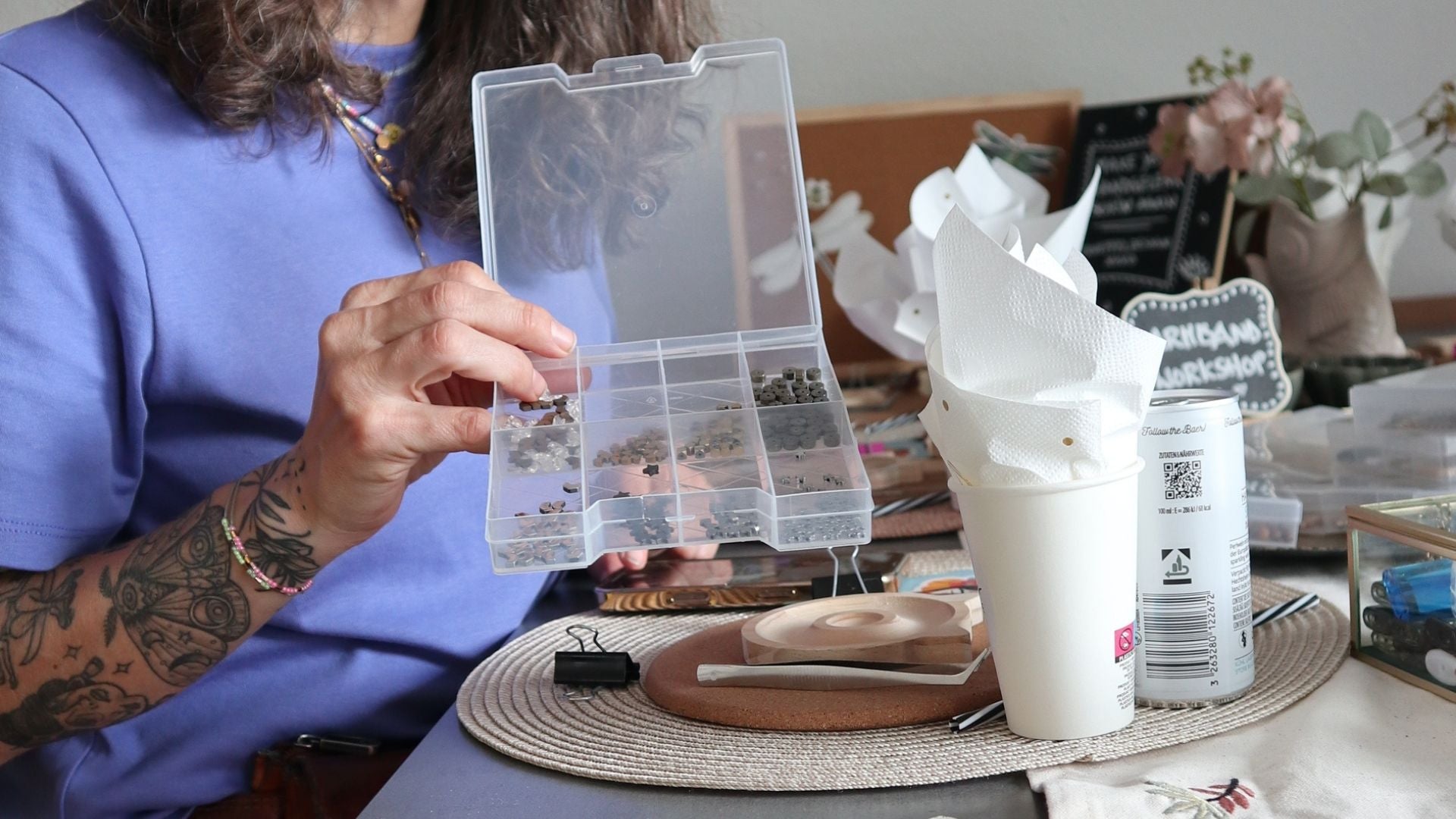 Frau mit Tattoos hält Perlen Sortierbox beim Armband Workshop, im Hintergrund Holztablett, Werkzeug und Tafel mit Aufschrift Armband Workshop